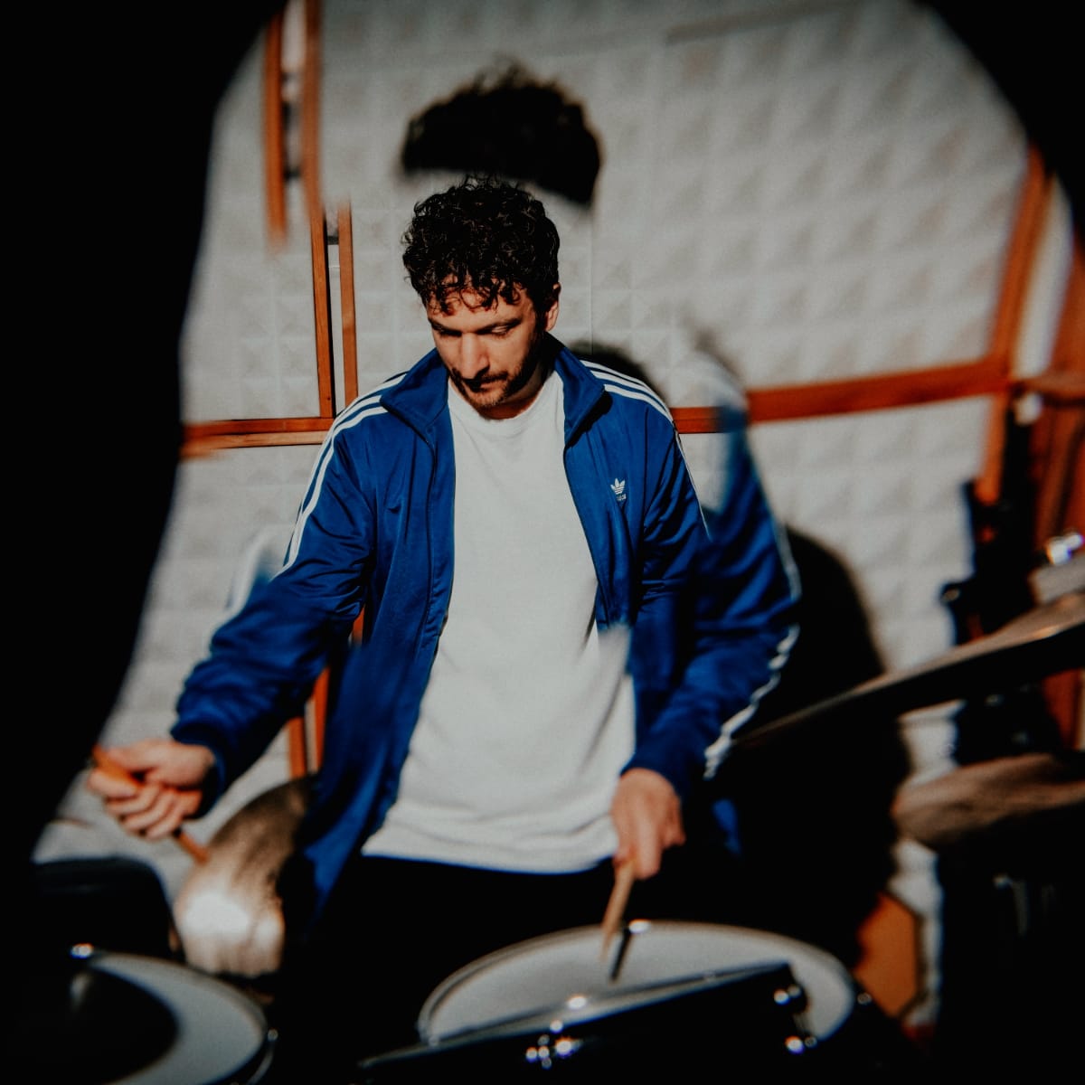 Peter Gall brings a stick down on a snare, head bowed, in a blue track jacket, his shadow thrown across the white pyramid acoustic foam behind him.