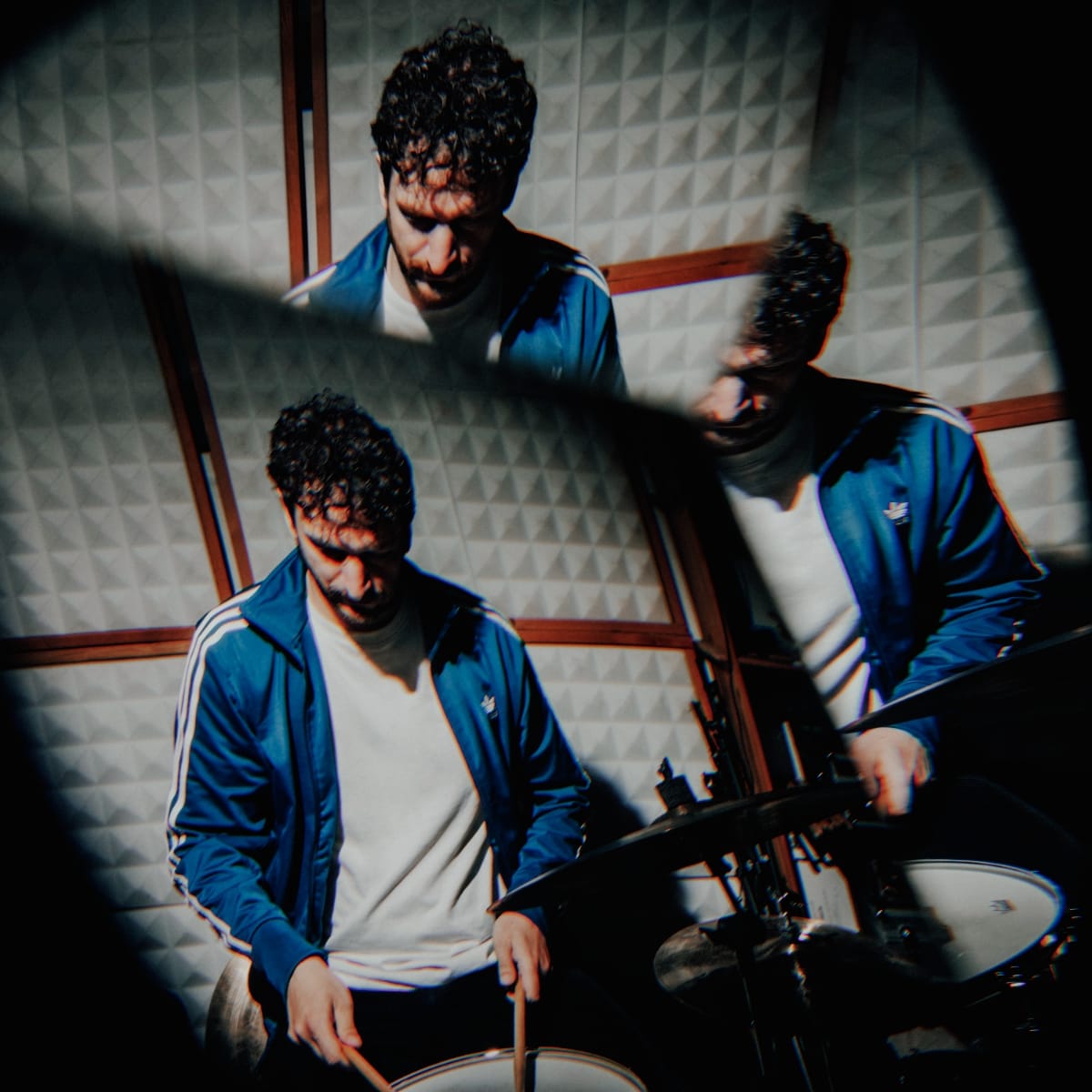 Peter Gall plays drums in a mirrored triple exposure, three overlapping views of him in a blue track jacket against a wall of white pyramid acoustic foam.