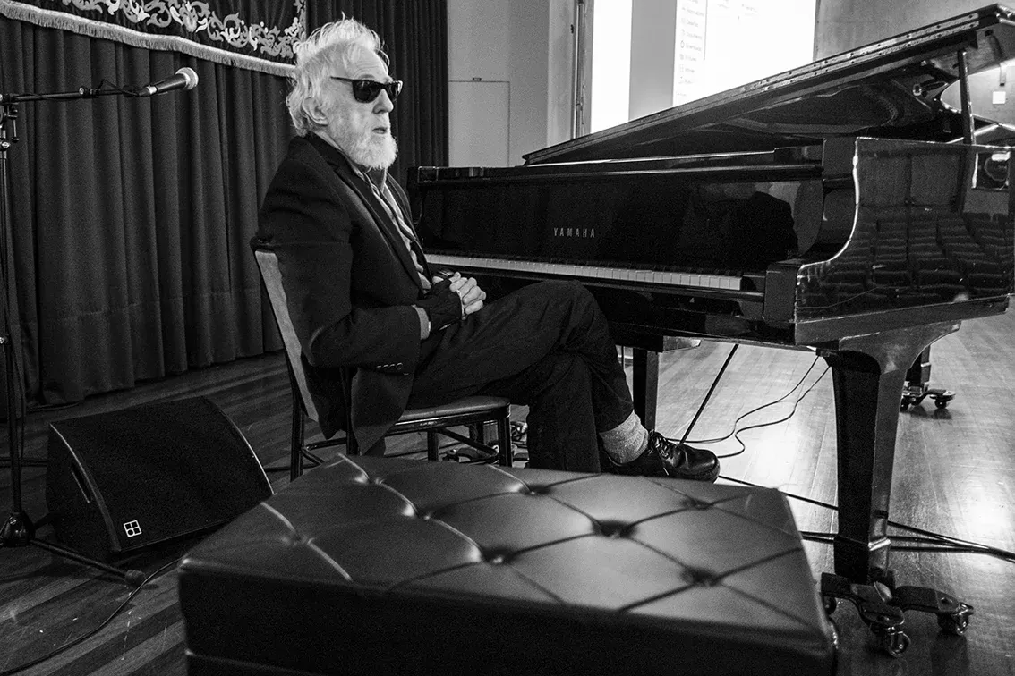 Ran Blake sits turned away from a Yamaha grand piano in sunglasses and a dark suit, legs crossed and hands clasped, on a stage with a curtain and floor monitor behind him.
