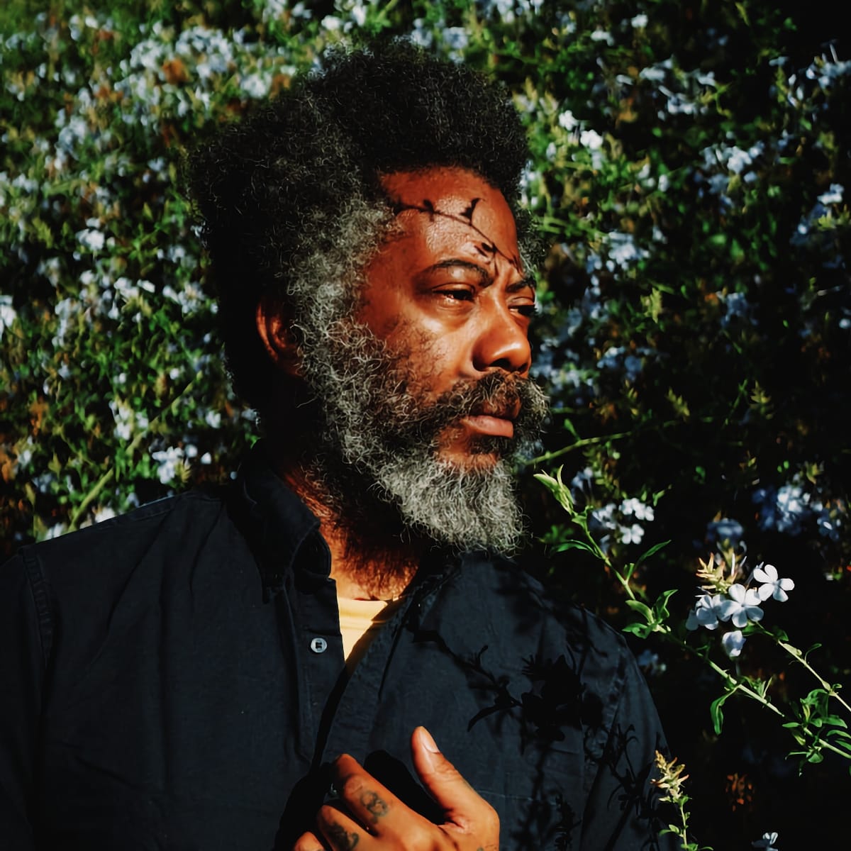 Robert Aiki Aubrey Lowe in profile against a wall of plumbago, pale blue flowers and dark leaves, low sun across his face, one tattooed hand raised to his chest.