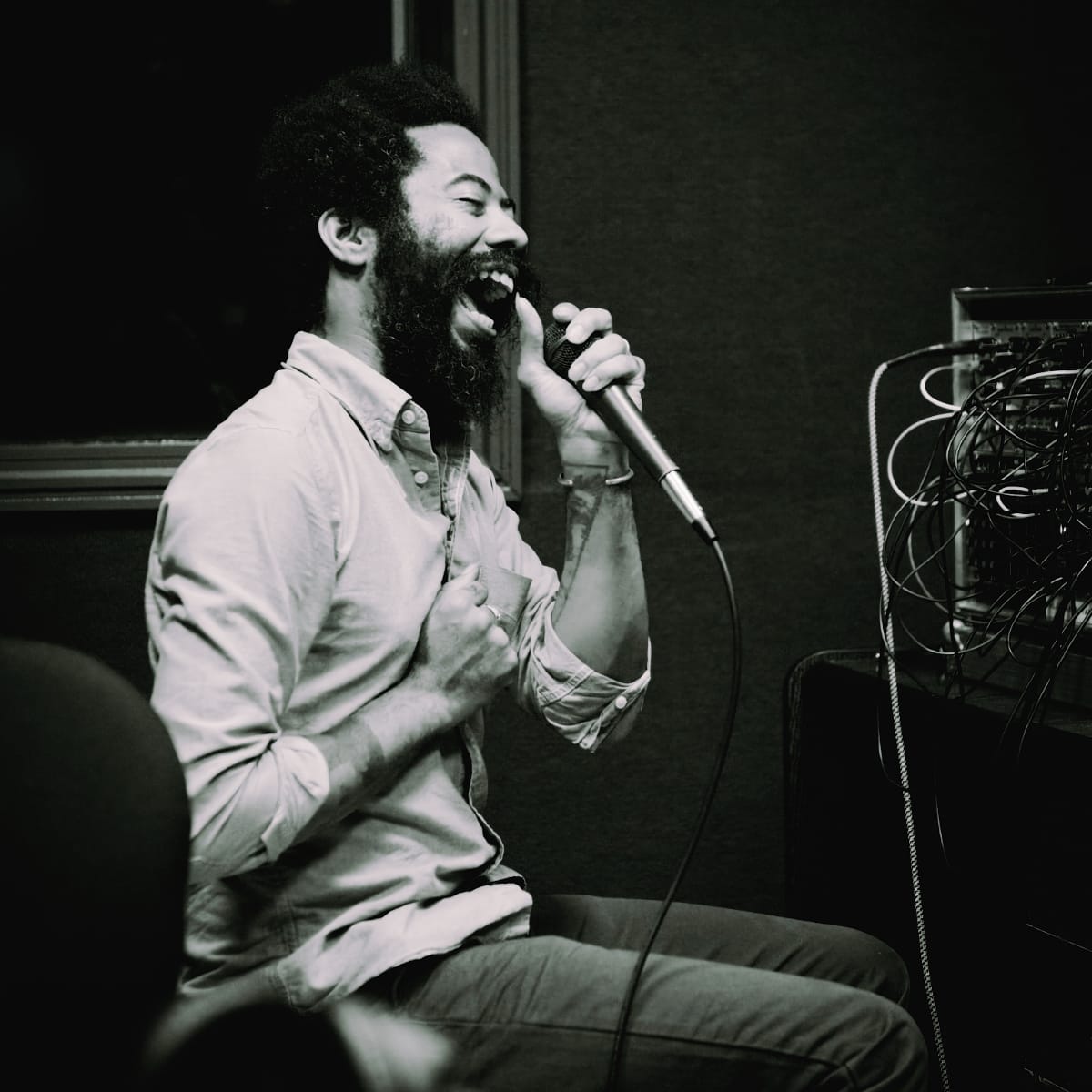 Robert Aiki Aubrey Lowe sings into a handheld microphone, seated with eyes shut and mouth wide, his other fist against his chest, a rack of tangled patch cables beside him. Black and white.