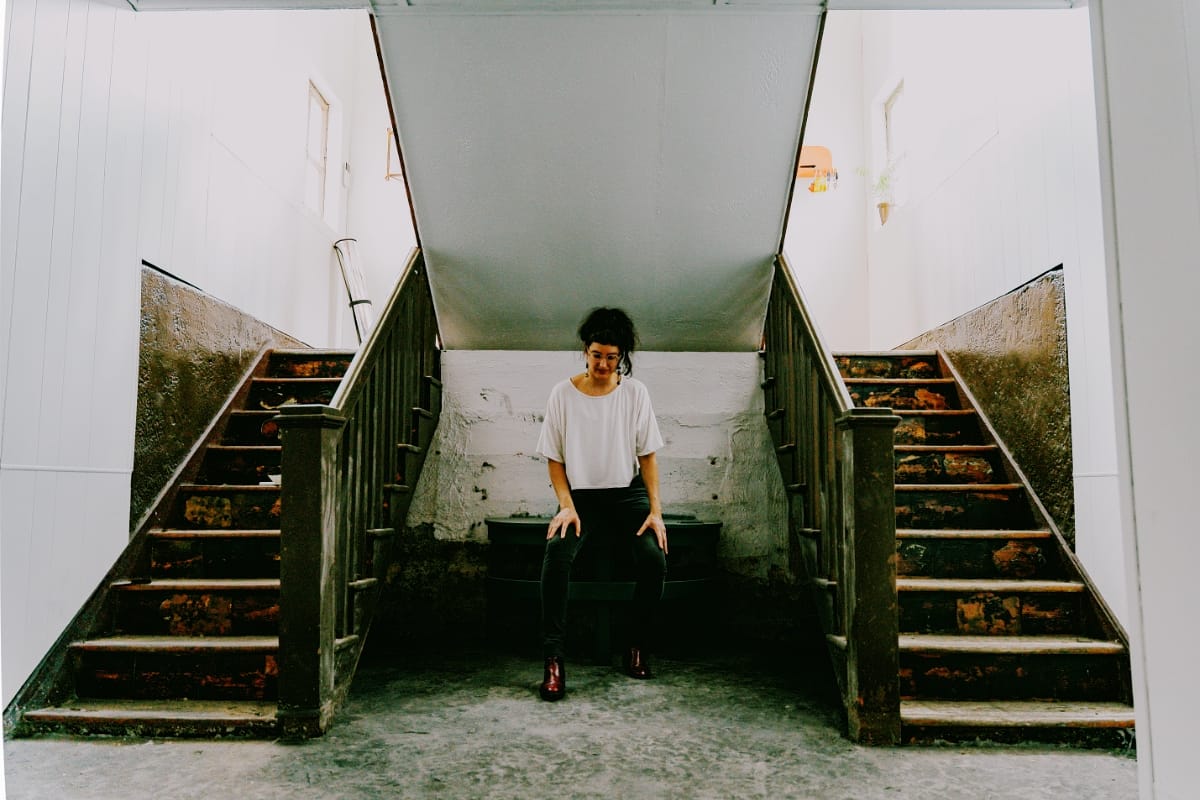 Sova sits on a low bench between two worn wooden staircases rising left and right in a white-walled stairwell, hands on her knees, looking down.