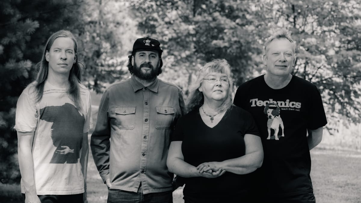 Black and white photo of Fred Thomas, Frank Rotondo, Windy Weber, and Carl Hultgren standing in a row on grass, evergreens and sunlit trees behind them.