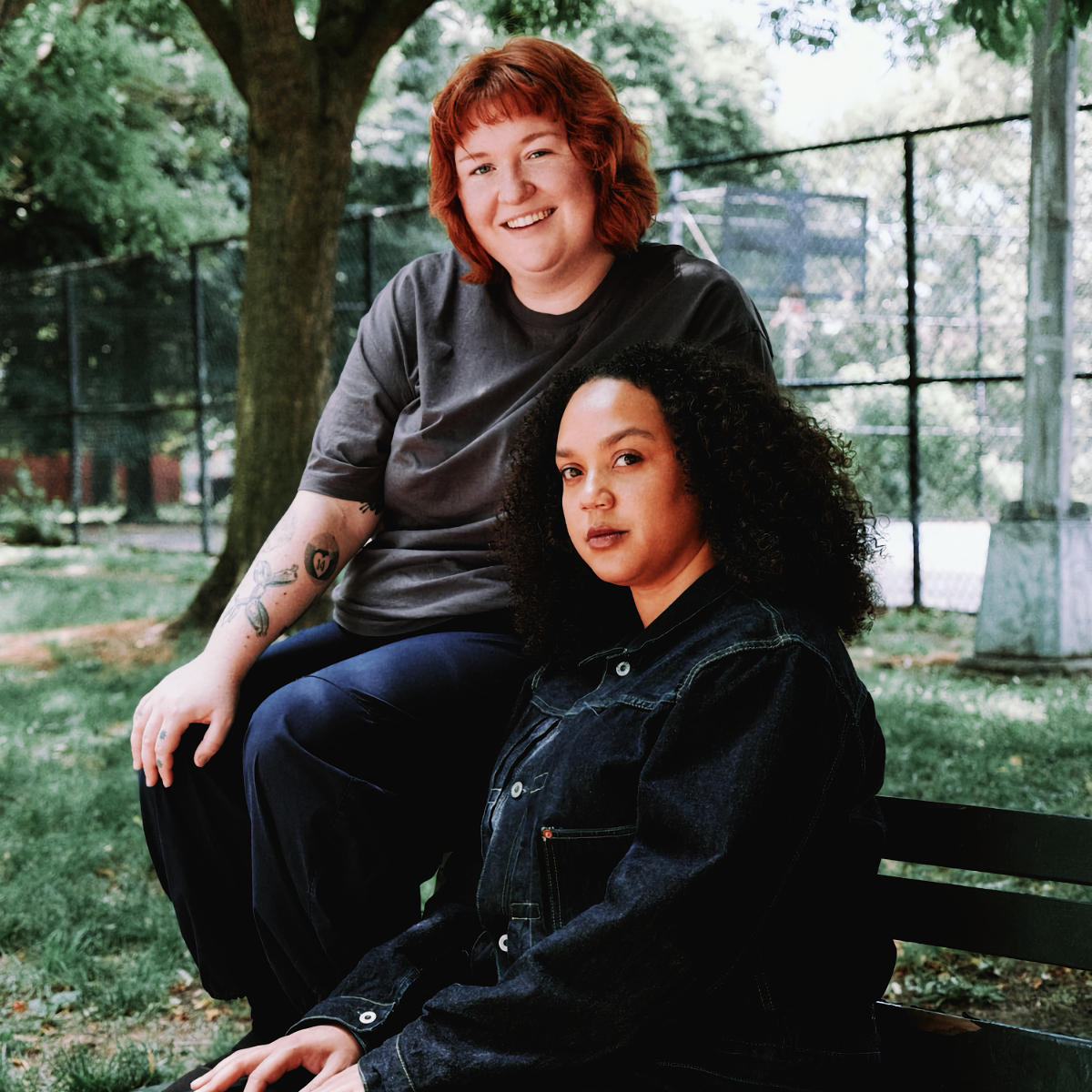 Nicole McCabe sits on the back of a park bench, smiling, behind Savannah Harris, who faces the camera in a dark denim jacket. A tree and a chain-link fence stand behind them.