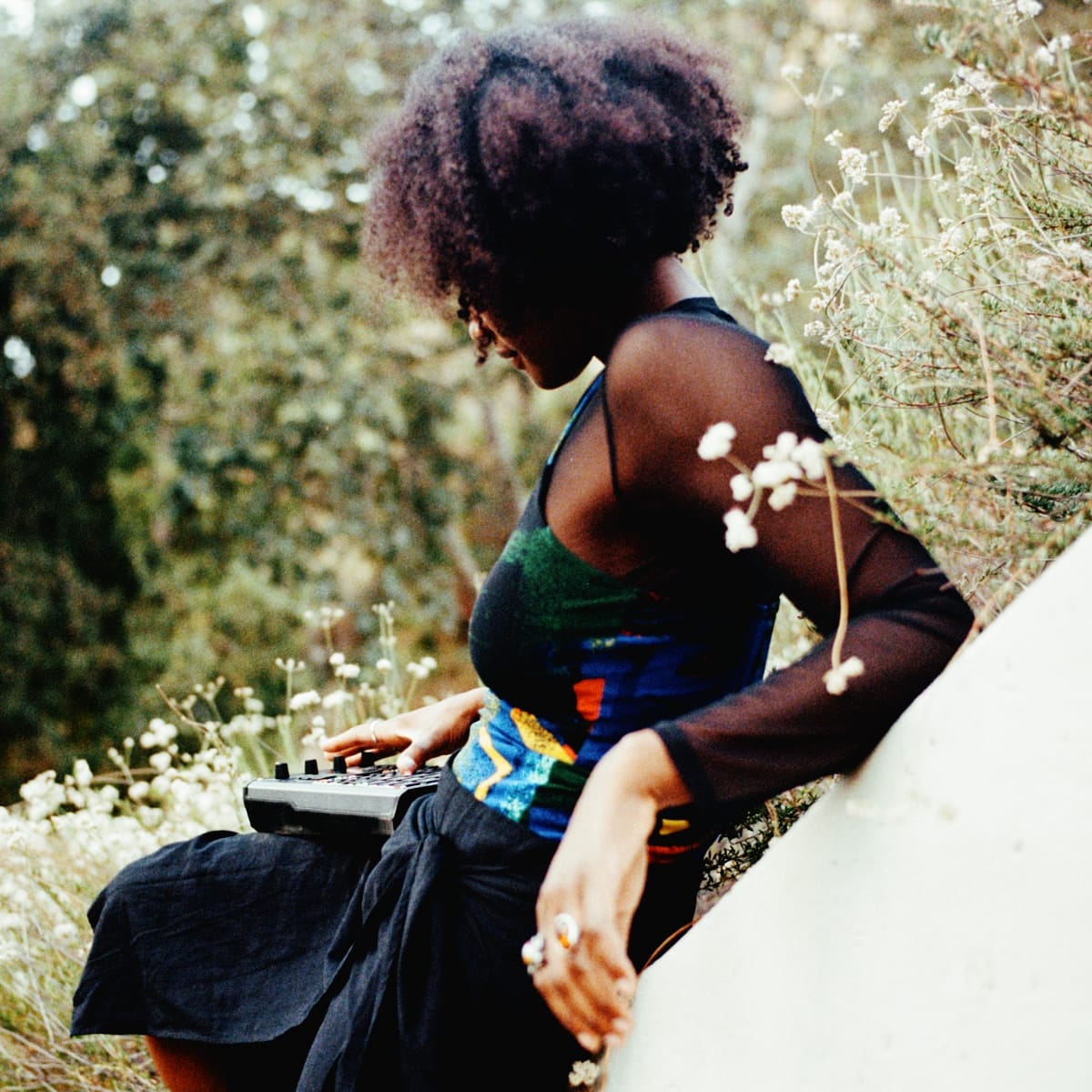 megiapa sits among white wildflowers, head bowed, one hand on a sampler balanced on her knee, the other arm draped over a pale concrete ledge.