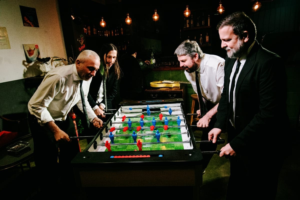 Members of Calibro 35 in white shirts and dark ties lean over a foosball table, mid-game, in a dim bar strung with bare Edison bulbs.