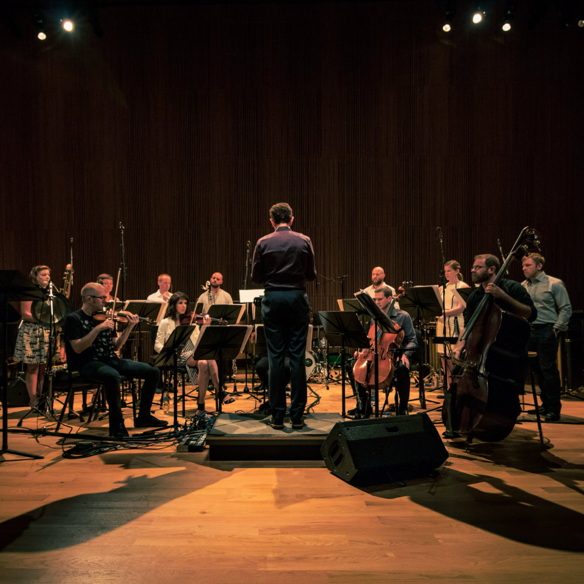 Alarm Will Sound rehearse in a semicircle on a wood-floored stage, a conductor on the podium with his back to the camera, strings, winds, and bass lit warm against a dark wall.