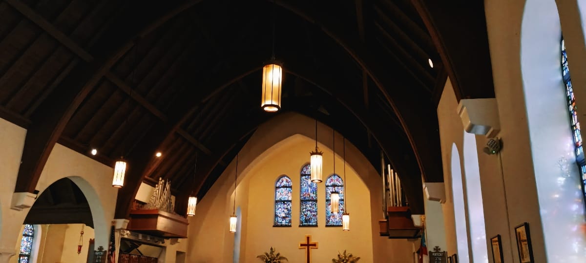 The nave of All Saints Episcopal Church, Winter Park, Florida: dark timber arches, hanging lanterns, organ pipes on both sides, and three stained glass windows above a wooden cross.
