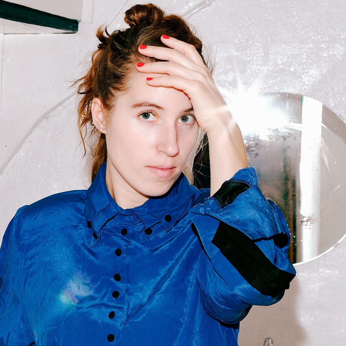 Charlotte Greve in a blue silk shirt, one hand pressed to her forehead, looking straight at the camera beside a round mirror flaring with light.