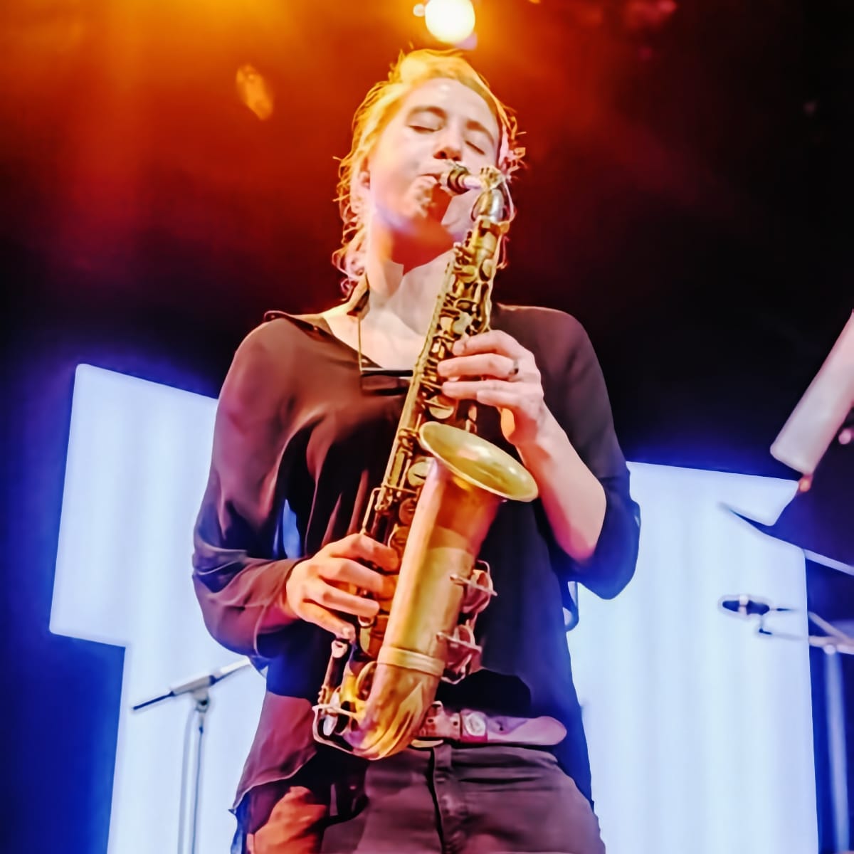 Charlotte Greve plays alto saxophone on a stage, eyes closed, lit in orange and blue against white panels.