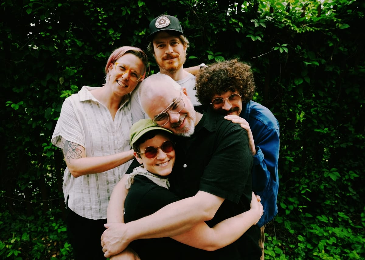 Crying Laughing pile into a group hug in front of a wall of dense green leaves, all five grinning at the camera.