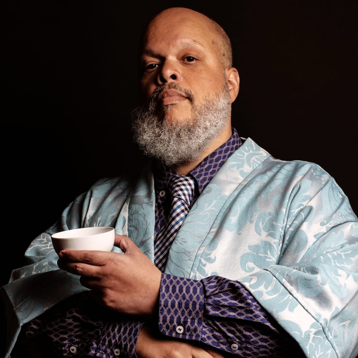 Ed Motta against a black background in a pale blue damask robe worn over a patterned navy shirt and checked tie, holding a small white cup and looking into the camera.