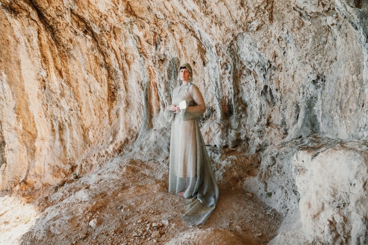 Jolanda Moletta stands barefoot in a shallow cave of ochre and gray rock, wearing a long sheer gray hooded gown, a small hand mirror held at her chest.