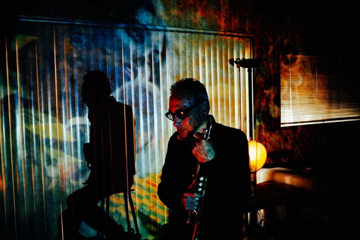 Jon Hassell in dark glasses holds his trumpet in a dim room, his silhouette and slatted light from vertical blinds projected across a wall of blue and orange imagery beside a glowing lamp.