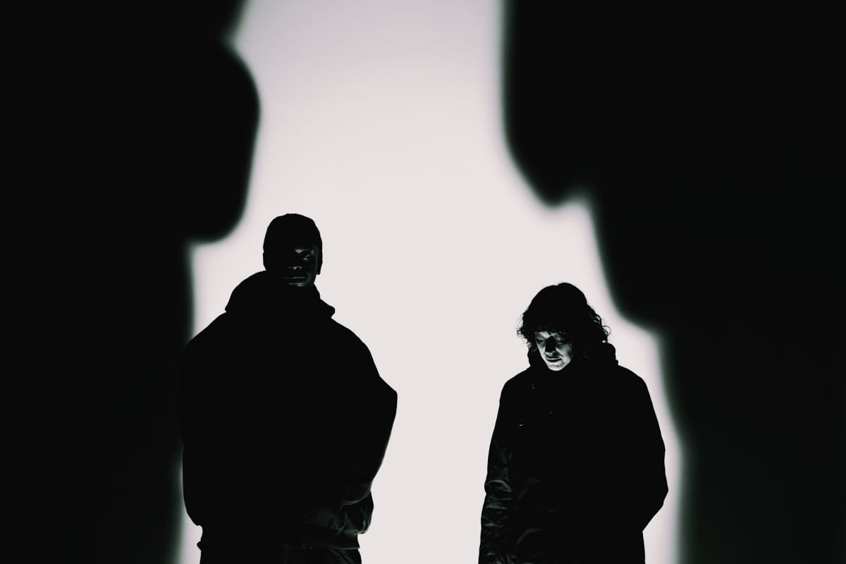 Chris Ryan Williams and Ka Baird stand backlit against a white wall, faces barely lit, Baird looking down, blurred dark shapes framing them in the foreground.