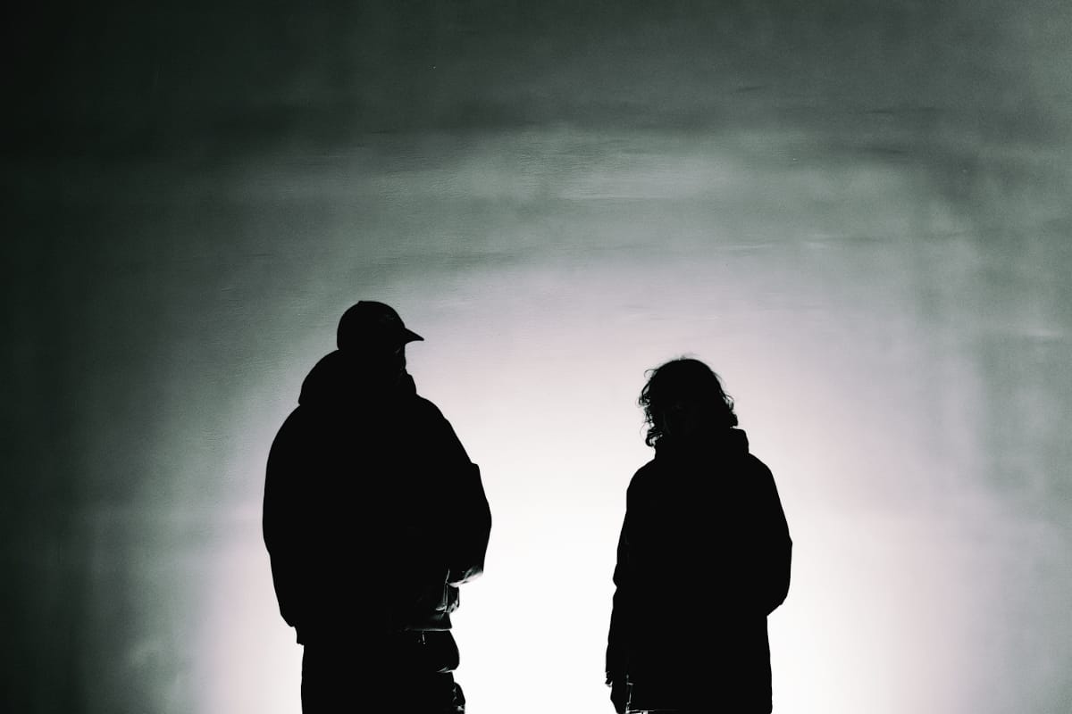 Chris Ryan Williams, in a cap, and Ka Baird stand in silhouette against a pale, misty backlit wall, Williams turned toward Baird.