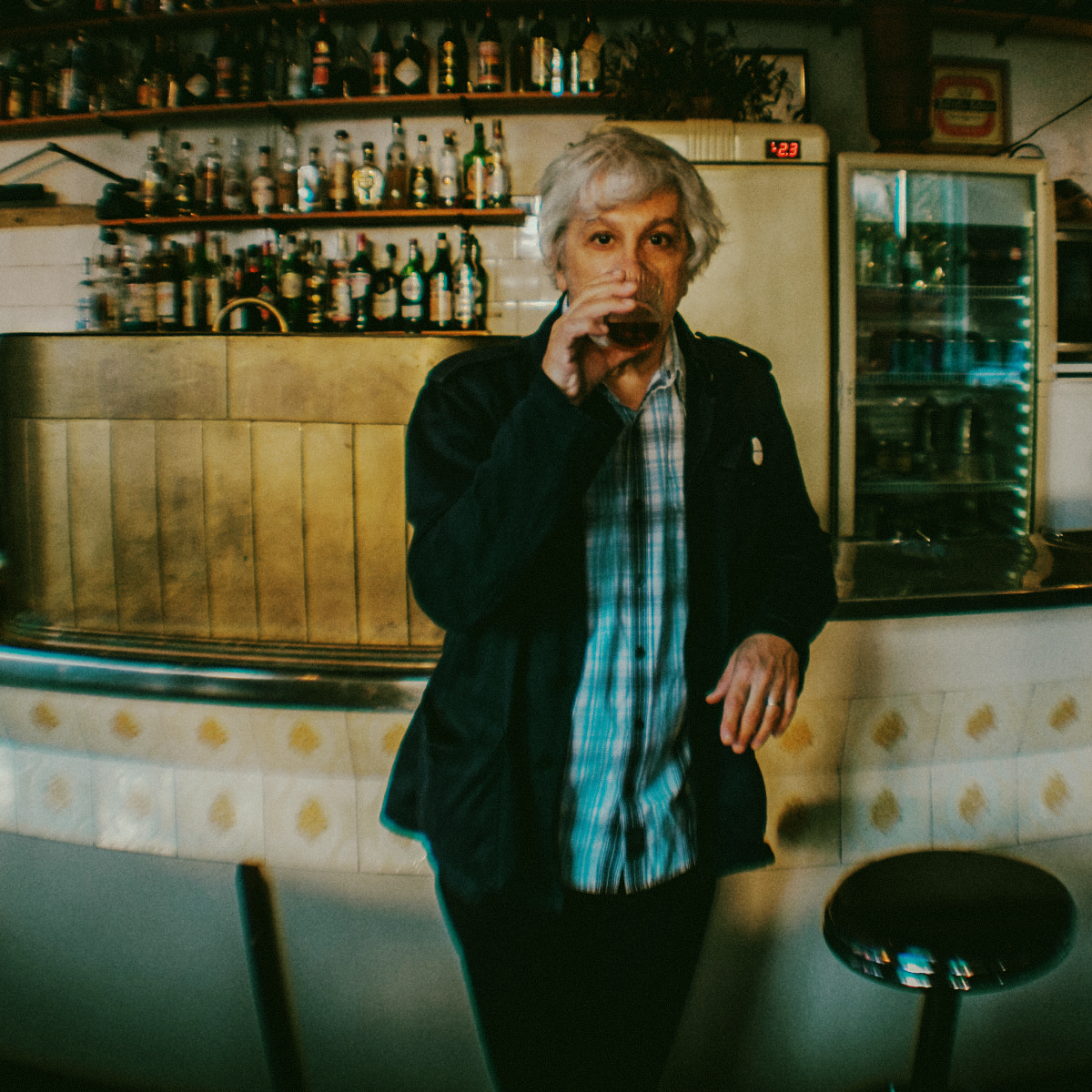 Lee Ranaldo sips from a glass at a tiled bar counter, dark jacket over a plaid shirt, shelves of liquor bottles and a glass-front fridge behind him.