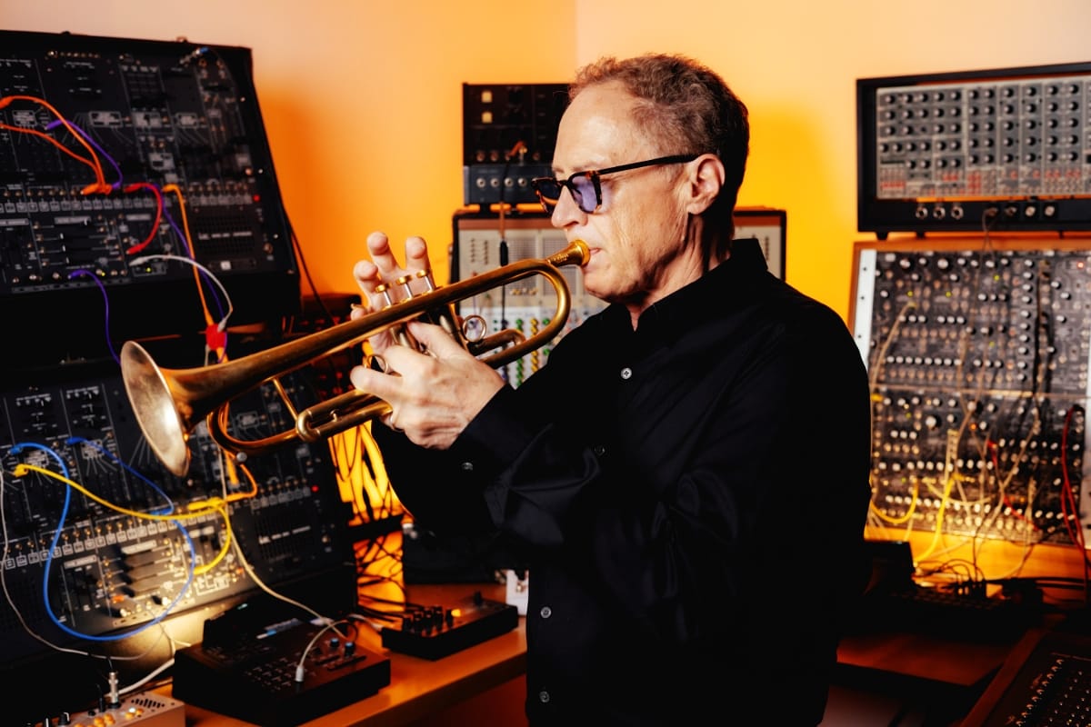 Mark Isham plays trumpet in tinted glasses, flanked by walls of patched modular synthesizers glowing in warm orange light.
