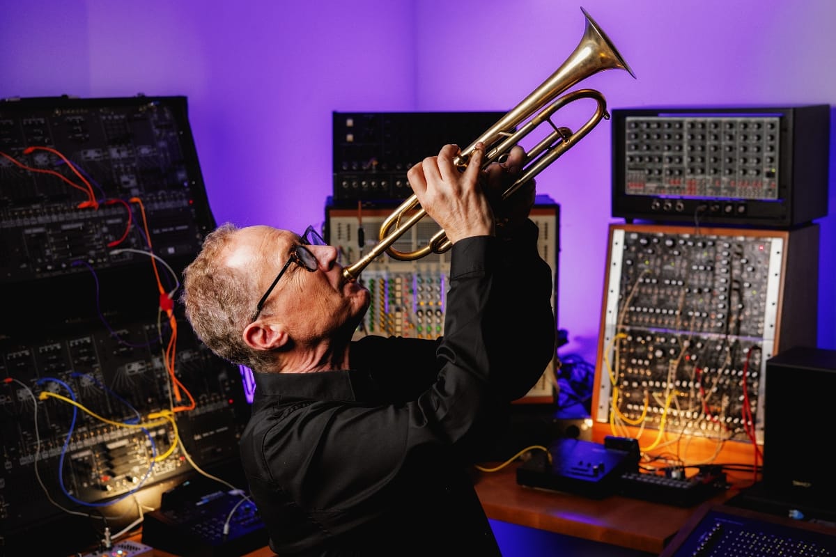 Mark Isham tilts his head back and points his trumpet upward, framed by modular synthesizers patched with colored cables under violet light.