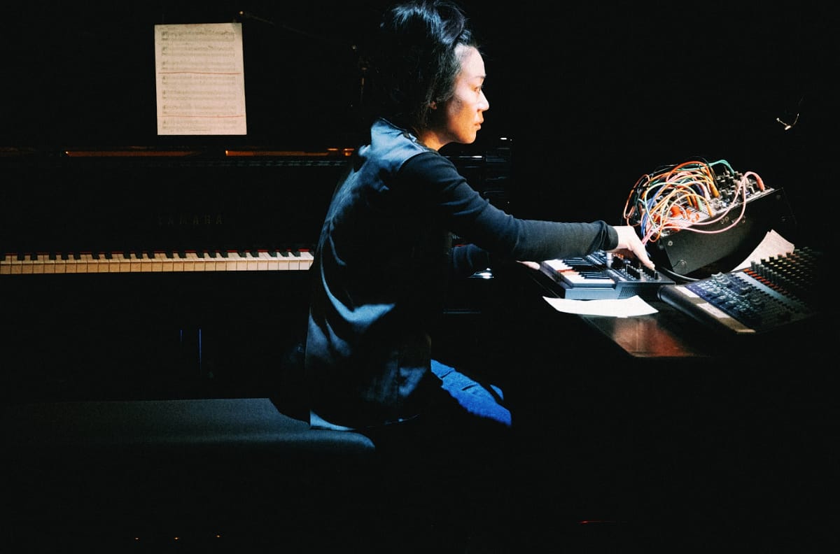 Midori Hirano performs The Juniper Tree at De Vooruit in Ghent, turning from a grand piano to a small synth and a modular rig strung with colored patch cables, under a single spotlight.