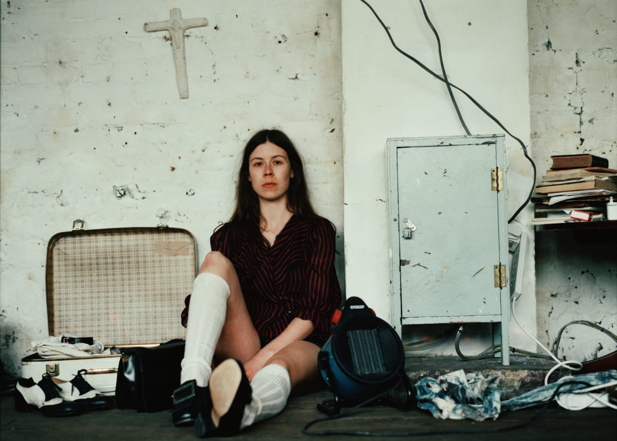 Nina Winder-Lind sits on the floor against a whitewashed brick wall beneath a plaster cross, in a striped dress and white knee socks, beside an open suitcase and a space heater.