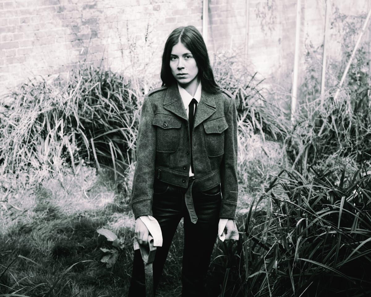 Nina Winder-Lind stands in an overgrown walled garden in a wool military-style jacket and black tie, one hand on a spade handle, gazing at the camera. Black and white.