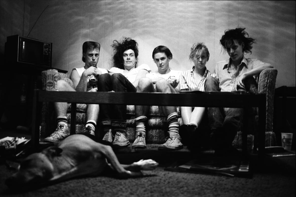 The Butthole Surfers sit shoulder to shoulder on a couch behind a low coffee table, staring into the camera, while their dog, Mark Farner, sleeps on the carpet in front.