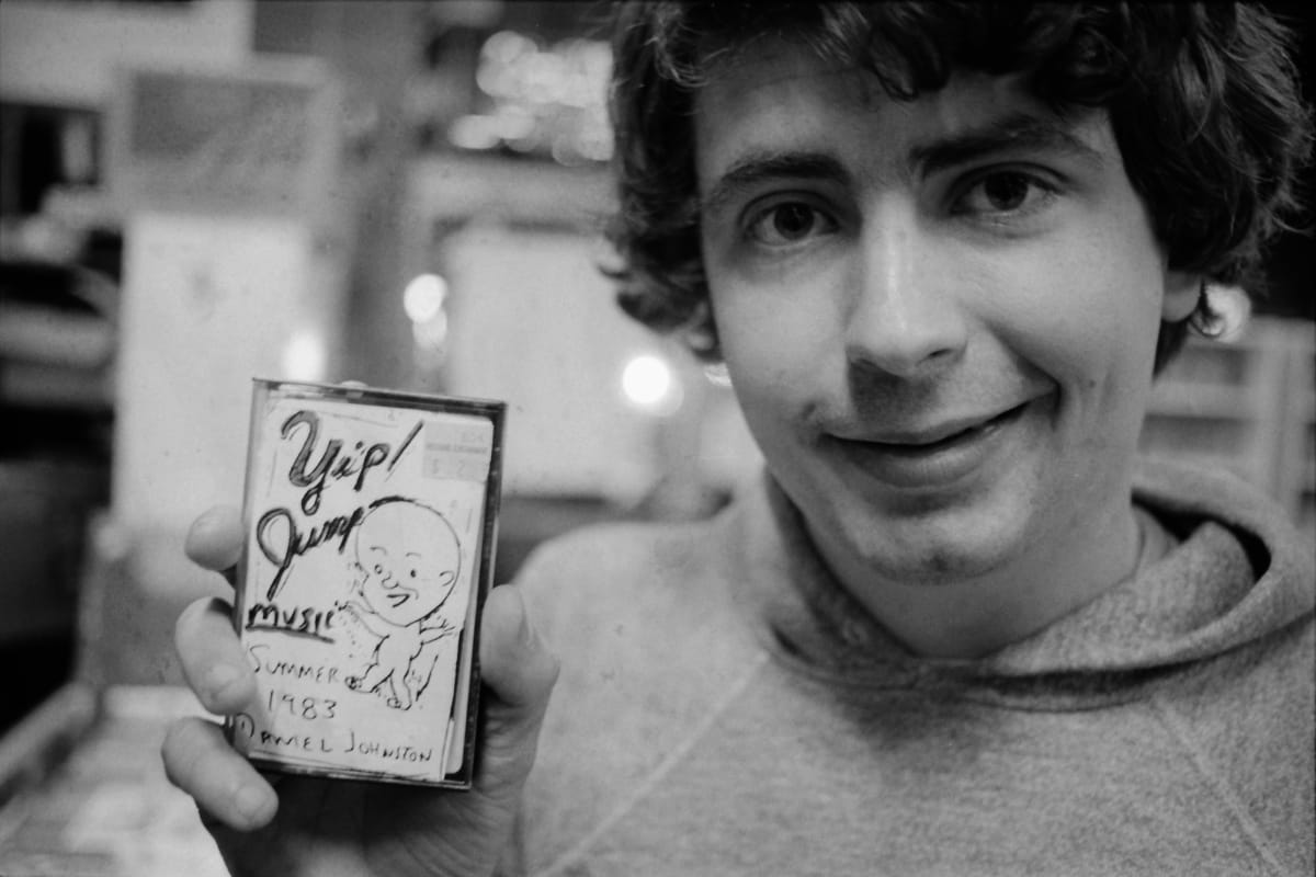Daniel Johnston grins at the camera in a record store, holding up his cassette Yip/Jump Music, its hand-drawn cover marked Summer 1983 above his name.