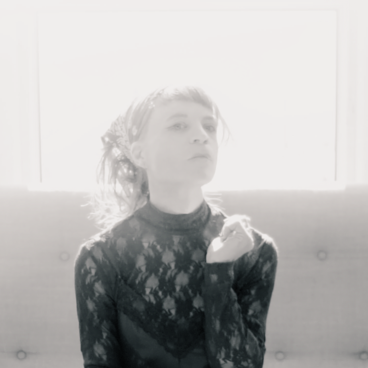 Washed-out black-and-white portrait of Agnes Haus in a dark lace top and patterned headscarf, one hand at their chest, seated on a sofa before a bright window.