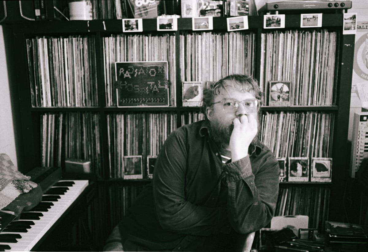 Sam Cook-Parrott, in glasses and a dark shirt, rests his chin on his fist before floor-to-ceiling record shelves, a Radiator Hospital sign among the LPs. Black and white.