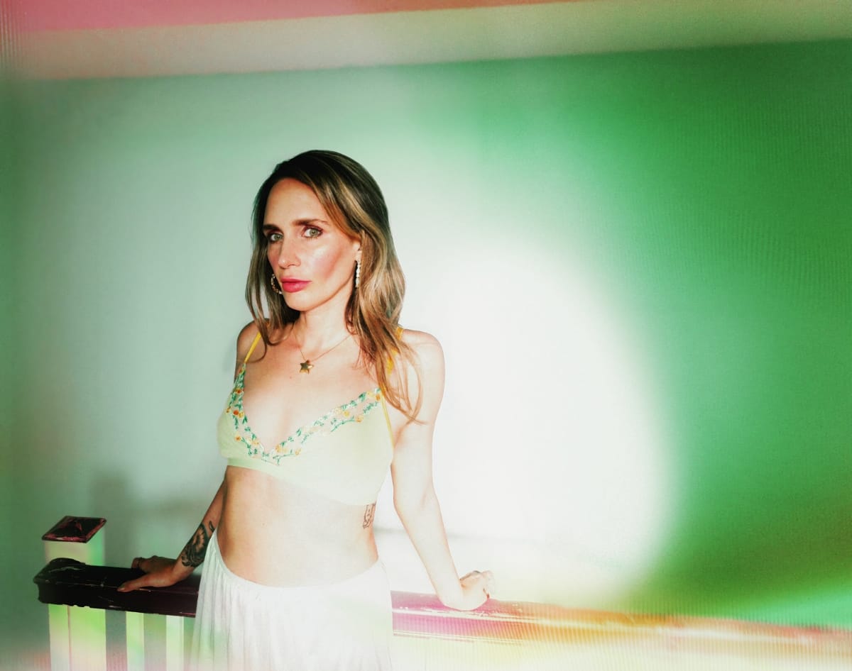 Sadie Dupuis leans on a wooden railing and meets the camera, in an embroidered yellow top and white skirt, against a hazy wash of green and white light