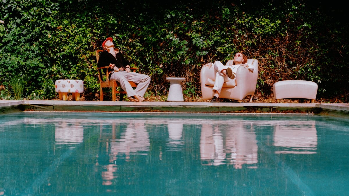 Sons of Sevilla lounge across a turquoise pool from the camera, one tipped back in a wooden chair in a red cap, the other sunk into a cream armchair in sunglasses, ivy rising behind them.