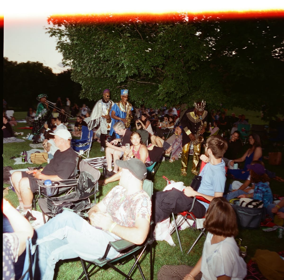 Sun Ra Arkestra horn players in robes and crowns walk among a lawn-chair audience at Storm King Art Center at dusk, under a broad tree, a streak of red film flare across the top.