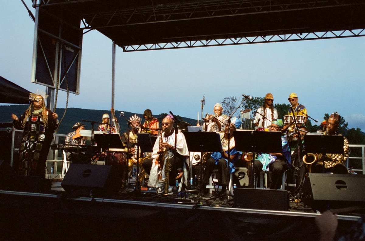 The full Sun Ra Arkestra in robes and headdresses plays an open-air stage at Storm King Art Center at dusk, a vocalist at left and wooded hills behind.