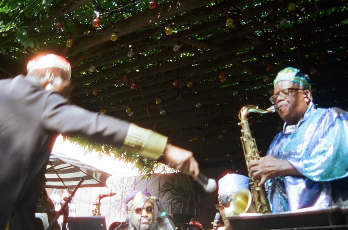 Bandleader Knoel Scott, seen from behind, holds a microphone out toward a Sun Ra Arkestra saxophonist in a shimmering blue robe, under Union Pool’s vine-covered pergola strung with colored bulbs.