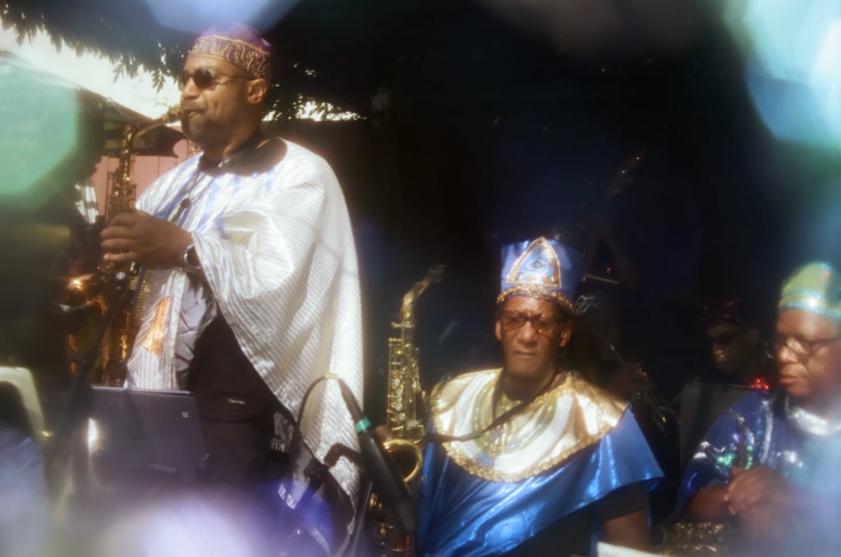 Sun Ra Arkestra saxophonists in a white striped cape and blue-and-gold robes perform at Union Pool, their caps and costumes catching the light through soft lens flares.