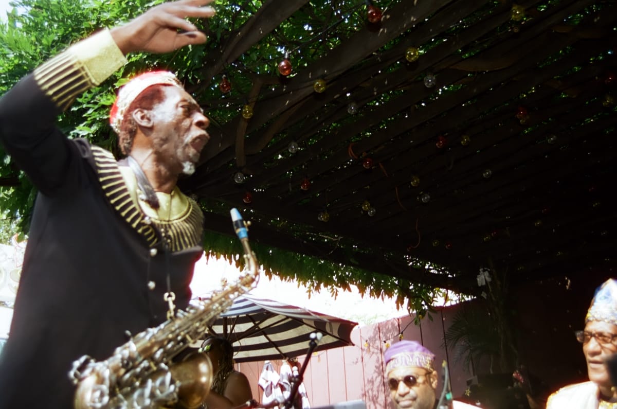 Knoel Scott, saxophone hanging from its strap, raises an arm mid-cry as he leads the Sun Ra Arkestra beneath Union Pool’s vine-covered pergola, bandmates in embroidered caps below.