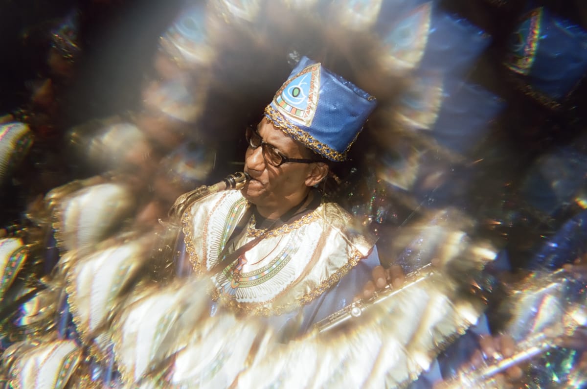 A Sun Ra Arkestra saxophonist in a blue cap with an eye emblem and a gold-trimmed Egyptian-style collar plays at Union Pool, a flute in hand, haloed by prism-lens reflections.