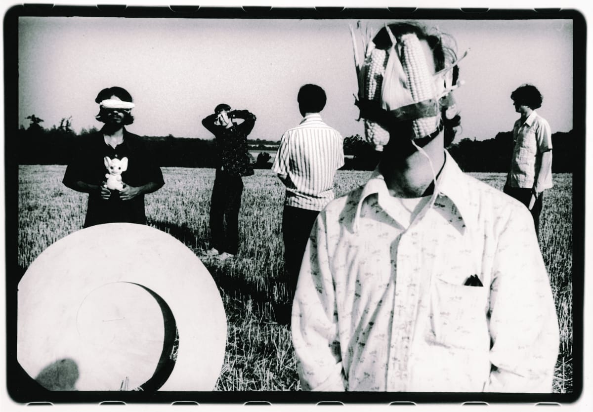 The Olivia Tremor Control in a stubble field, in black and white: one member’s face masked with ears of corn, another blindfolded holding a toy elephant, two turned away.