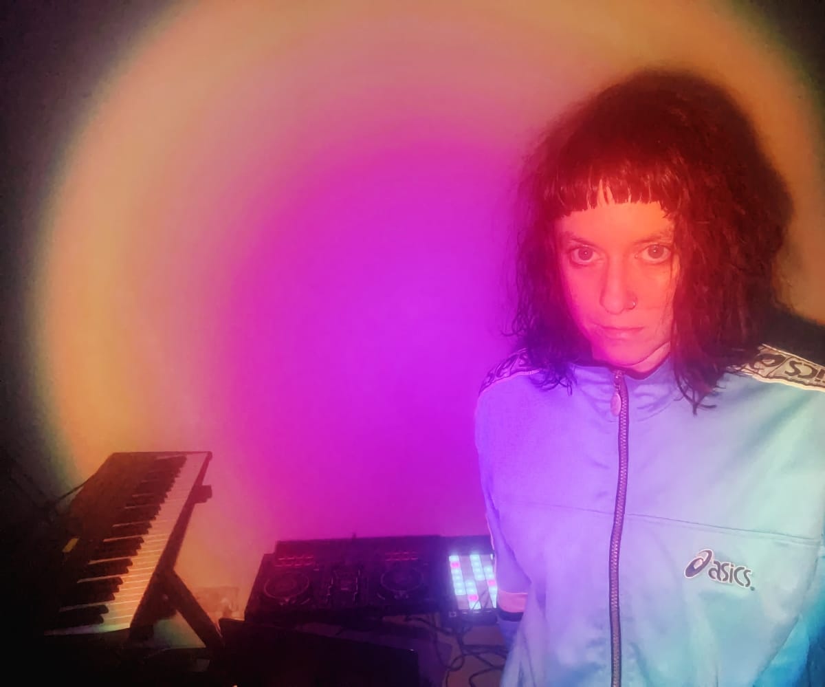 Tiiva stands in a home studio washed in a magenta and amber halo of light, a keyboard and a lit-up controller behind them, gazing straight at the camera.