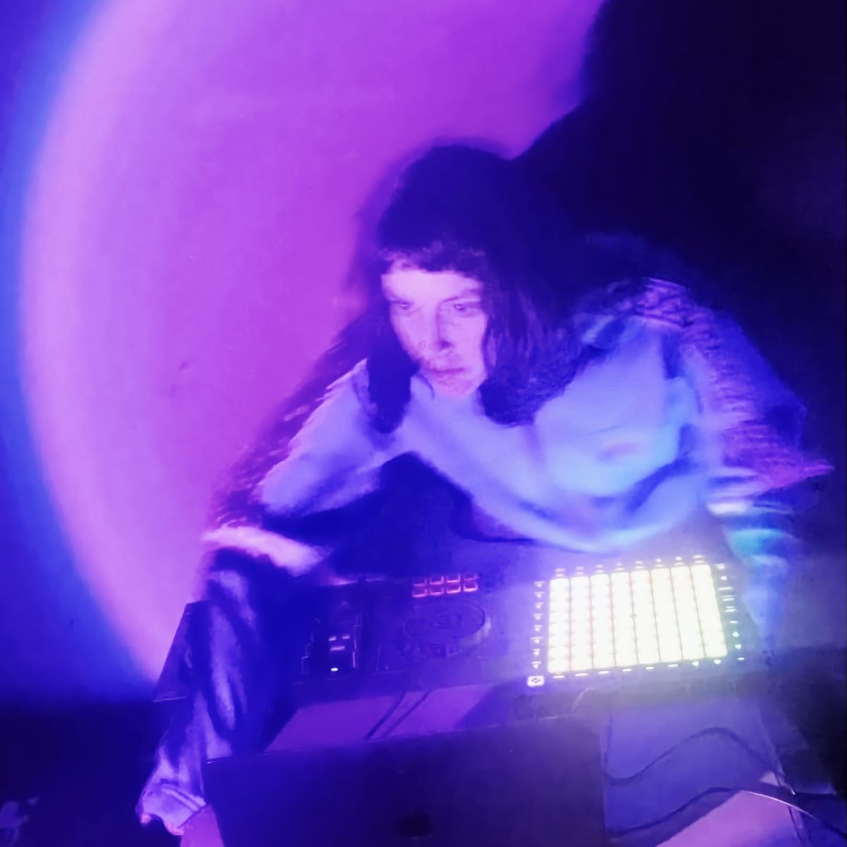 Tiiva leans over a DJ controller and a glowing grid of pads, the shot motion-blurred in violet and blue light.