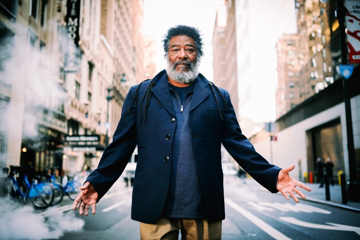 Wadada Leo Smith stands in the middle of a city street with his arms held out and palms open, wearing a navy jacket, as steam rises past parked bikes behind him.