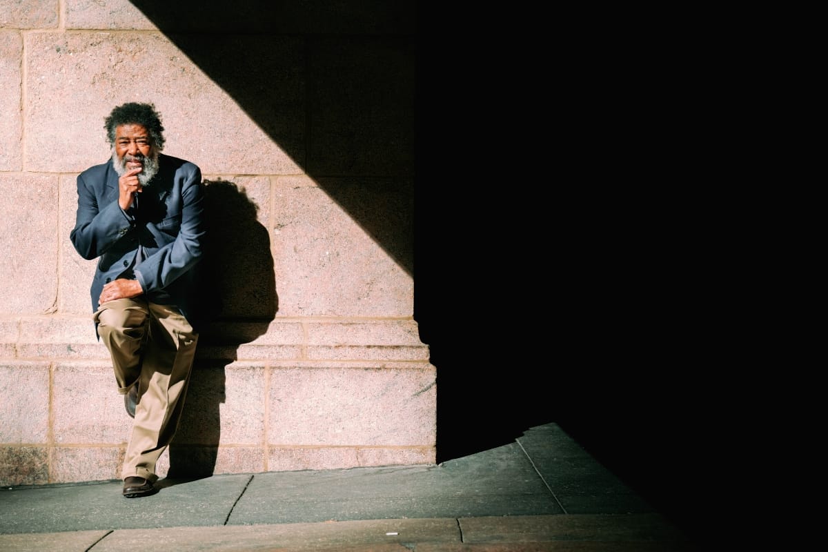Wadada Leo Smith leans against a sunlit granite wall in a navy jacket and khakis, one foot propped behind him and a hand at his chin, beside a deep band of shadow.