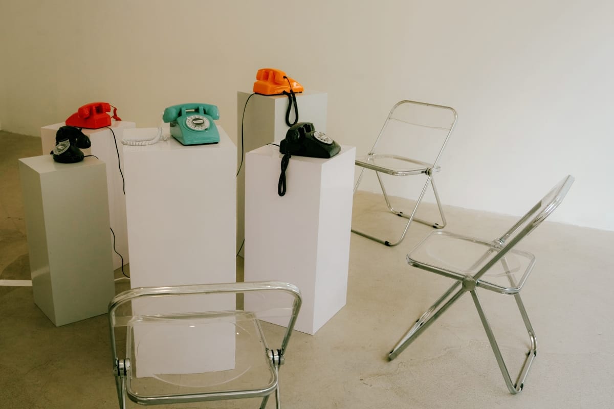 Five rotary telephones in red, black, teal, and orange sit on white plinths in a bare gallery, clear folding chairs set around them for listeners.