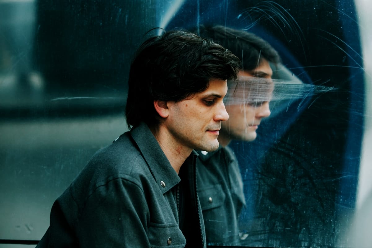 Brett Bullion of deep glens in profile, looking down beside a scratched blue-black panel that mirrors his face in a smeared reflection.