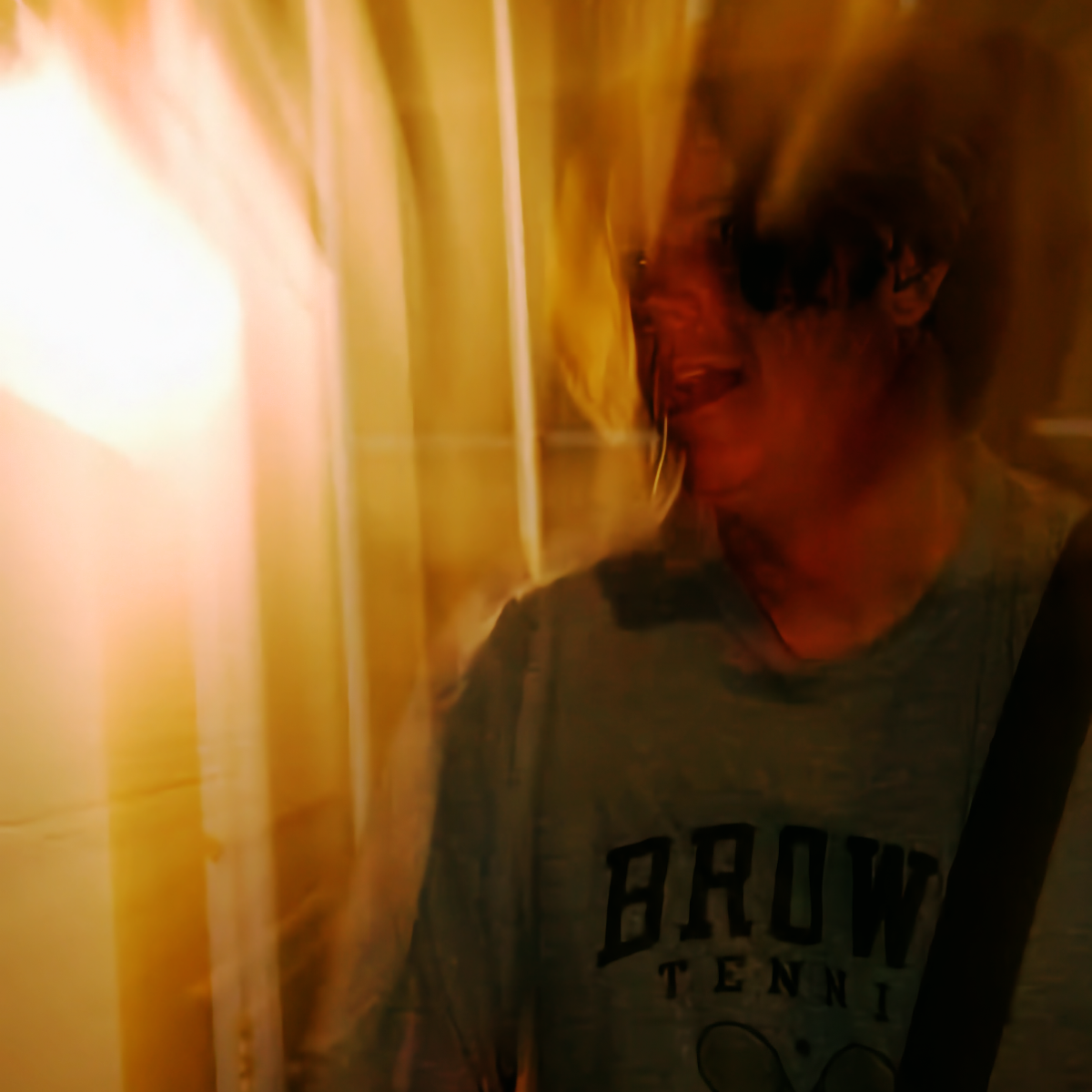 John Andrew Fredrick of the black watch, face blurred mid-motion and mouth open, wearing a gray Brown Tennis T-shirt and washed in amber and orange light.