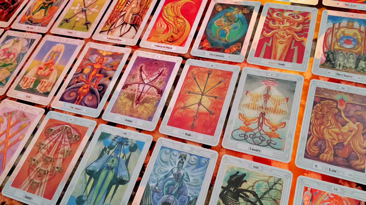 Rows of Thoth tarot cards spread across a fiery red cloth, their vivid painted images including The Magus, The Chariot, Lust’s lion, and Death’s skeleton.