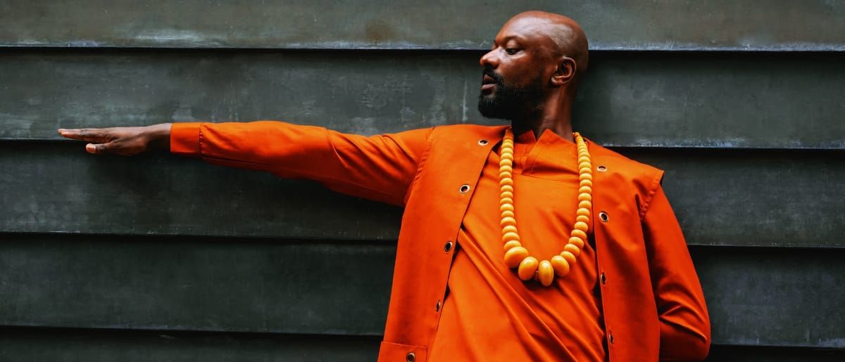Justin Hicks in bright orange jacket with wooden bead necklace, arm extended against dark slatted wall background. Photo by Malik Well.