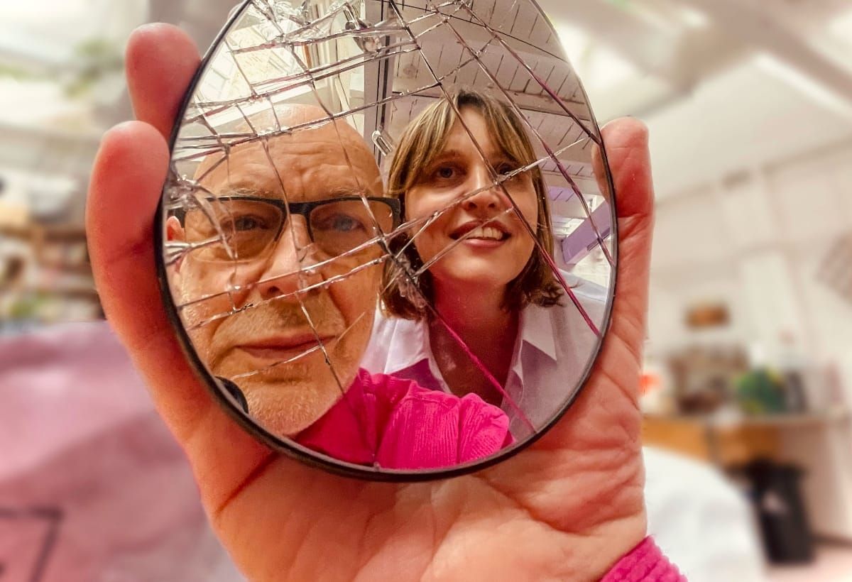 Brian Eno and Bette A reflected in handheld decorative mirror with geometric pattern, bright pink clothing visible. Photo courtesy of Brian Eno and Bette A.
