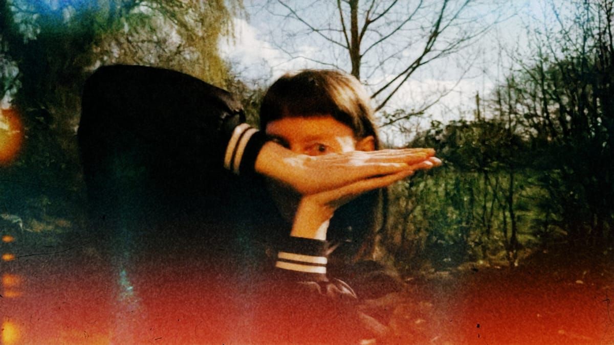 Loula Yorke with hand obscuring face in dreamy double exposure against bare trees, warm orange-red and cool blue-green tones.